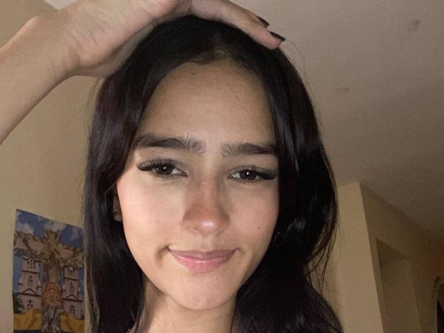 MelodyMansson has dark, wavy hair, a hand on her head, and is smiling with her pink lips slightly puckered. She has thick eyebrows and long eyelashes.