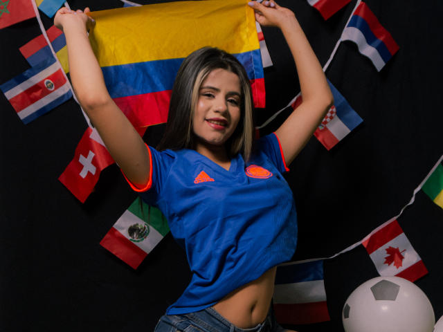SheylaSpooner with blonde hair, blue sports shirt, and jeans, holding a flag, smiling, arms raised, soccer ball on the right, flags in the background.