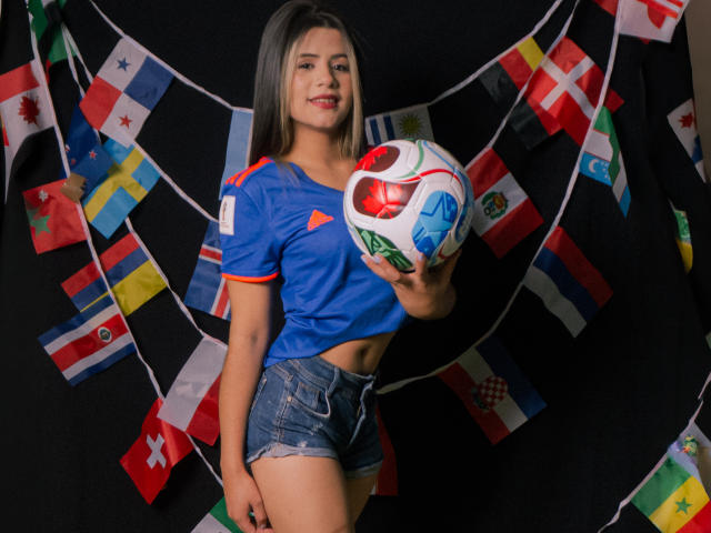 SheylaSpooner with long brown hair, blue soccer shirt, and denim shorts, holding a colorful soccer ball, smiling, black background with international flags. Boobs not visible, ass not visible.