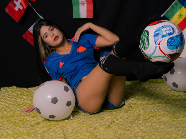 SheylaSpooner with long black hair, blue soccer shirt, red circle, black knee socks, denim shorts, and a soccer ball, legs up, lying on a green rug, red flag.