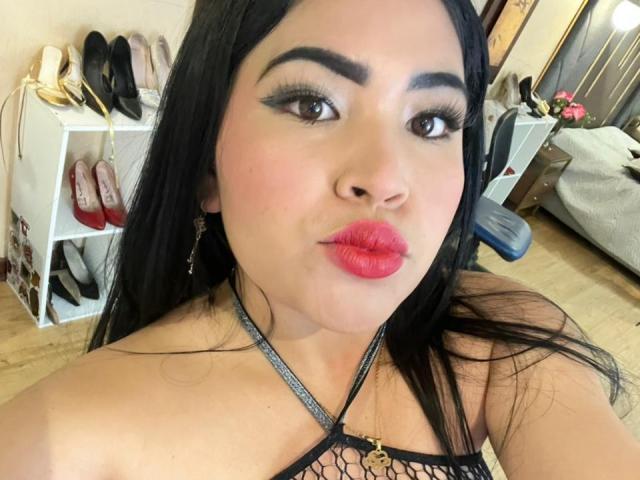 OliviGrace with long black hair, thick eyebrows, pink lipstick, and a black mesh top showing her tits, looking at the camera with a pouty expression.