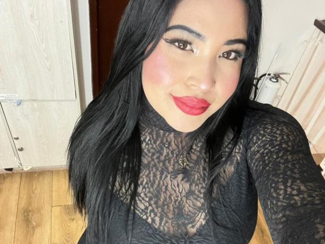OliviGrace with long black hair, red lipstick, and lace top, showing off her tits through sheer fabric, smiling at the camera. Wooden floor and door in background.