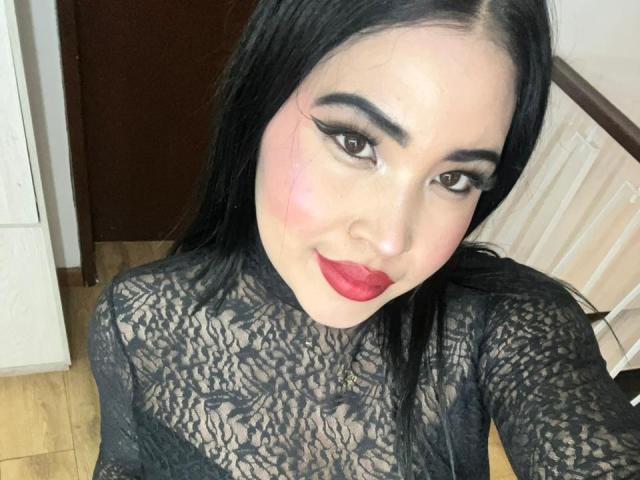 OliviGrace with long black hair, wearing a black lace top, has red lipstick, thick eyeliner, and a slight pinkish bruise on her right cheek. Her tits are not visible.