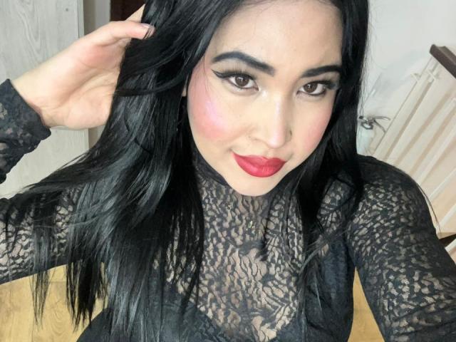 OliviGrace with long black hair, wearing a black lace top, red lipstick, and eyeliner, smiling at the camera, hand in hair, wooden floor, white wall, radiator in background.