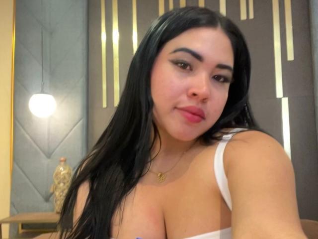 OliviGrace with long black hair, white top, showing her big boobs, pink lips, and subtle makeup, wearing a gold necklace, looking at the camera with a slight smile.