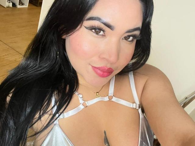 OliviGrace with long black hair, brown eyes, and pink lipstick, wearing a silver strappy top, showing her tits, and a small gold necklace, smiling at the camera.