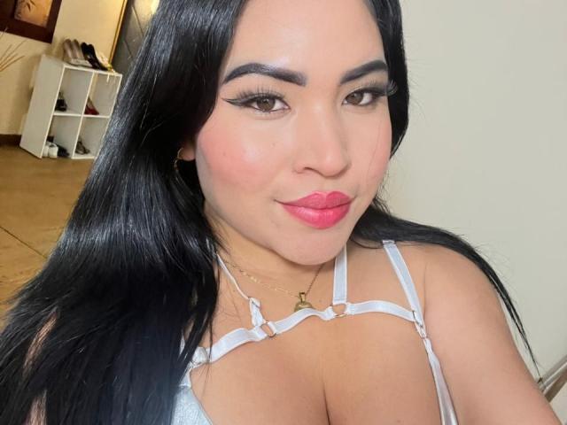 OliviGrace with long black hair, wearing a white strappy top, showing her large tits, pink lipstick, and a subtle smile, standing in a well-lit room.