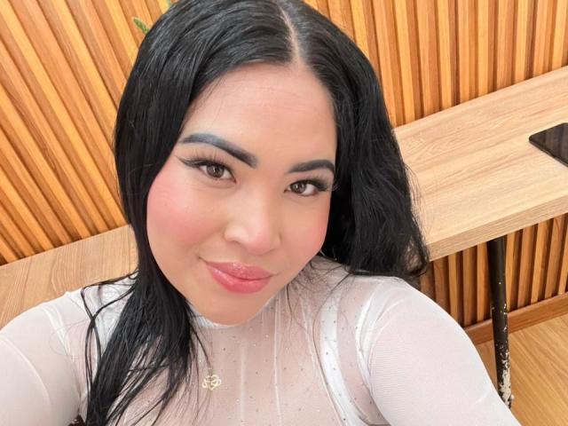 OliviGrace with black hair, brown eyes, and pink lips, wearing a sheer white top, showing her boobs, and a gold necklace, smiling at the camera.