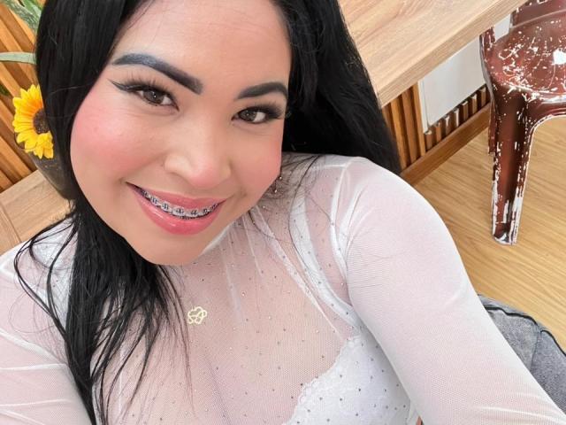 OliviGrace with black hair, pink lipstick, and white sheer top, smiling at the camera, showing metal braces on her teeth, wearing a gold necklace, sitting at a wooden table.