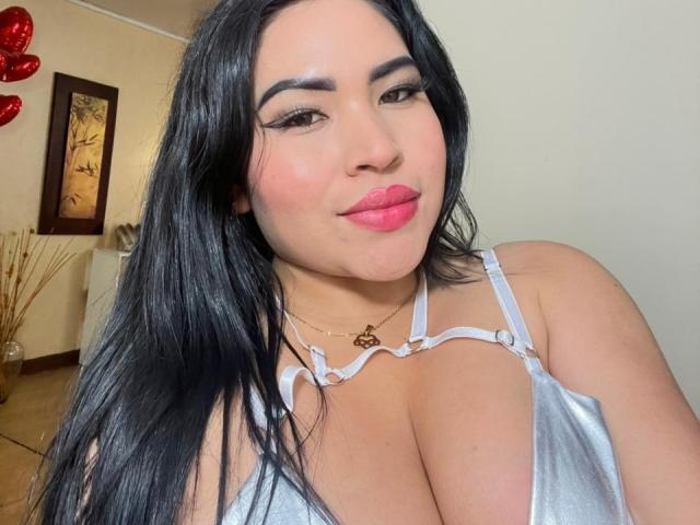 OliviGrace with black hair, pink lips, and a silver top showing her big tits, wearing a gold necklace, looking at the camera, smiling.