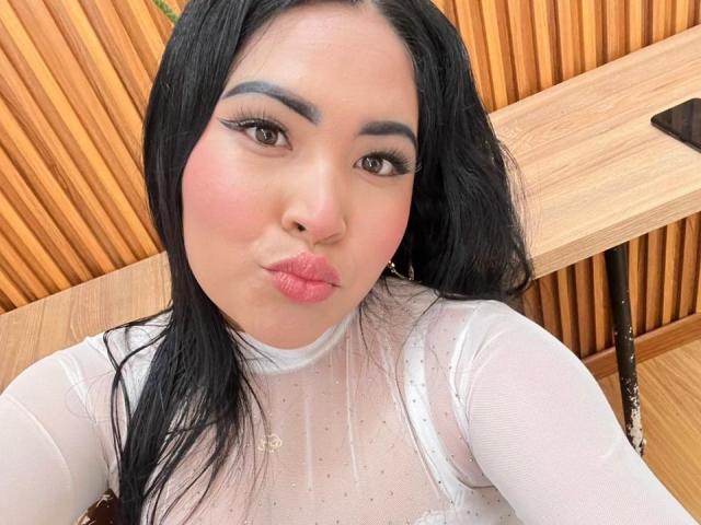 OliviGrace with black hair, pink lips, and white sheer top, pouting. She has bold eyeliner, pink blush, and small gold necklace. Her tits are visible through the top.