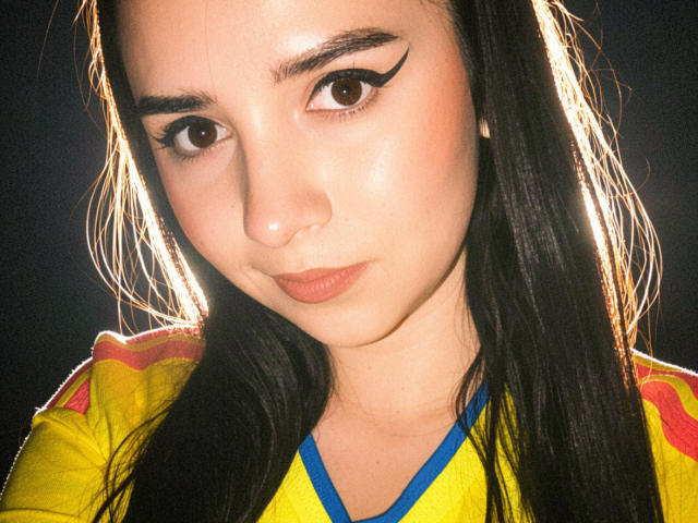 Kkatalina has pale skin, brown eyes with thick black eyeliner, and long black hair. She's wearing a yellow shirt with red accents and blue trim, looking down.