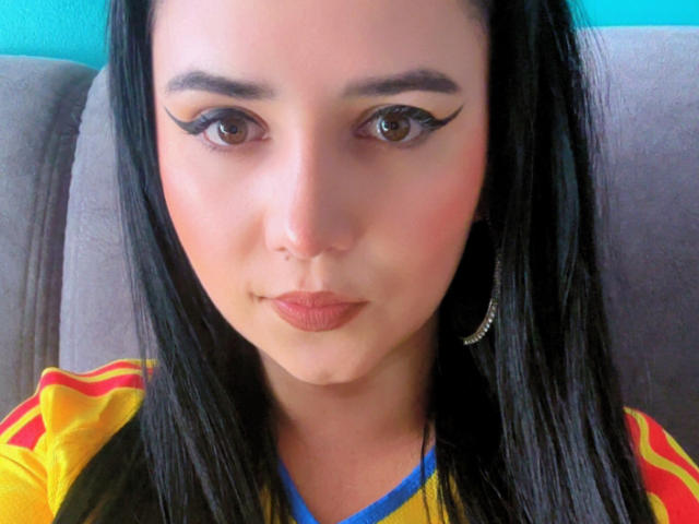 Kkatalina has black eyeliner wings, brown eyes, and is wearing a yellow and red shirt with a blue neckline. She has hoop earrings and is looking at the camera.