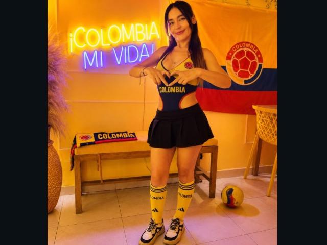 MindyLove in a Colombia soccer outfit, holding her tits, wearing a black skirt, yellow socks, and sneakers, standing with a soccer ball and neon "Colombia Mi Vidá" sign.