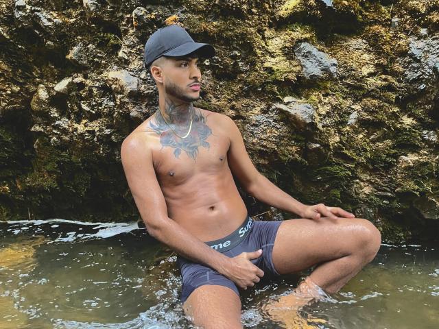 TheImperialMen, shirtless, dark skin, baseball cap, tattoo on chest, wearing dark blue shorts, sitting in shallow water, hand on bulge, rock wall in background, looking to the side.
