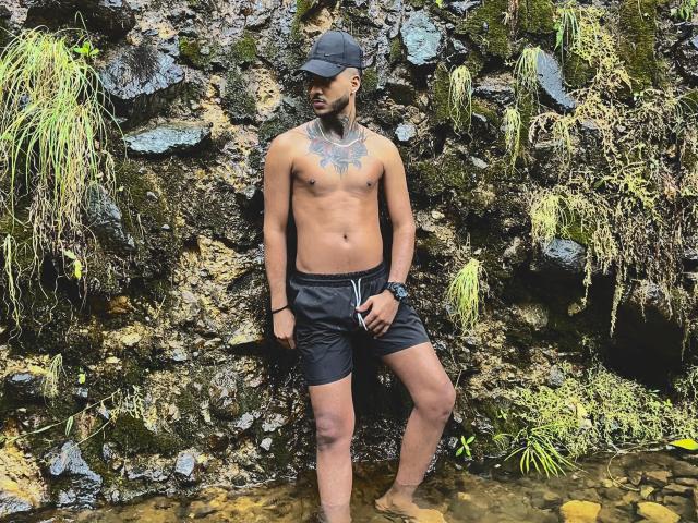 TheImperialMen stands shirtless against a rocky wall, wearing black shorts and a cap, with a tattoo on his chest, water at his feet, and green moss.
