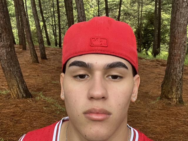 TheImperialMen has a red cap, brown eyes, thick eyebrows, light skin, small earrings, and a serious face, wearing a red shirt in a pine forest.