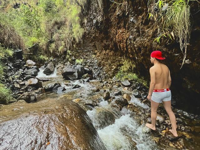 TheImperialMen, bare back, wearing red cap and grey shorts, standing in rocky stream, water flowing over rocks, greenery and moss-covered rocks in background, looking down at stream.