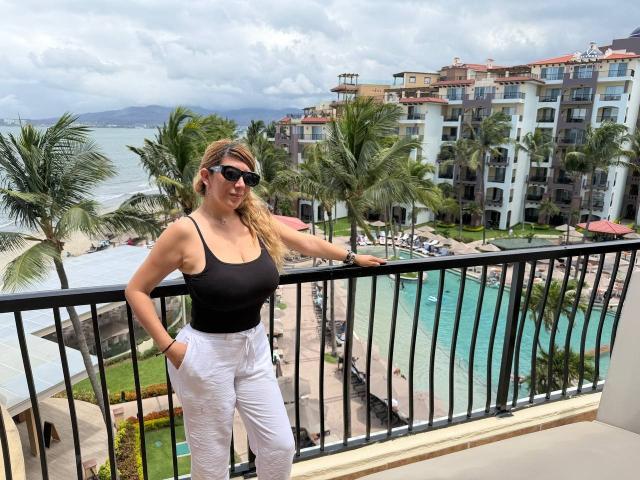 HotPamelaSex with blonde hair, black tank top, white pants, sunglasses, standing on balcony, hands in pockets, looking at camera, ocean and palm trees in background, cloudy sky.