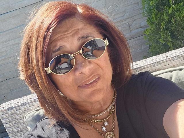 ClassybutNaughty with red-brown hair, wearing gold round sunglasses and multiple necklaces, smiling at the camera, showing a wrinkled face and wearing a black top.