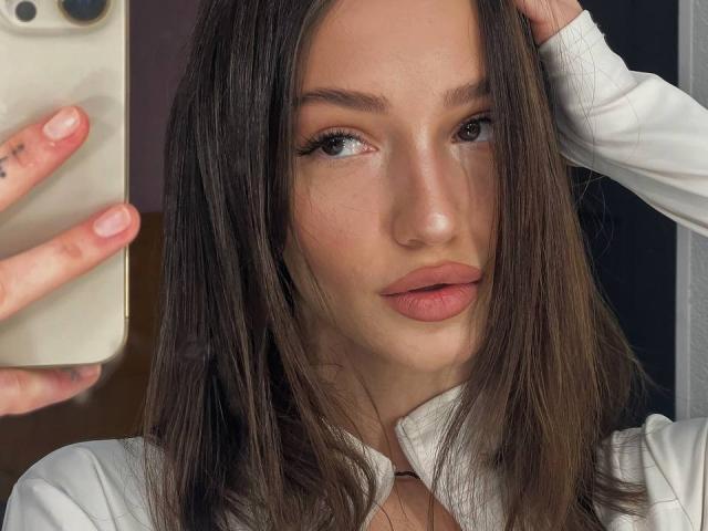 SophieKitty with long brown hair, pink lips, and white shirt, holding a phone in her left hand, looking at the camera with a subtle, sexy expression.