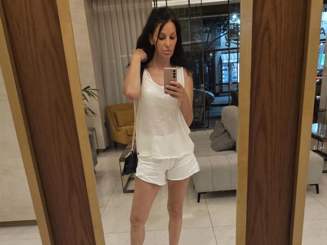JoliDanielle with long dark hair, wearing a white tank top and shorts, holding a phone, standing in front of a mirror, showing her tits and ass.