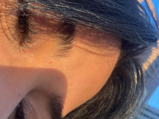 MajoJones's eye and upper cheek, with black eyelashes, brown eyelid, and short brown hair, in bright sunlight. Her skin is slightly shiny, showing natural texture.