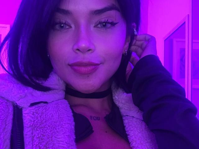 MajoJones with purple-tinted skin, black choker, fluffy white jacket, dark hair, and subtle smile, touching ear, showing small tits and tattoo under chin.