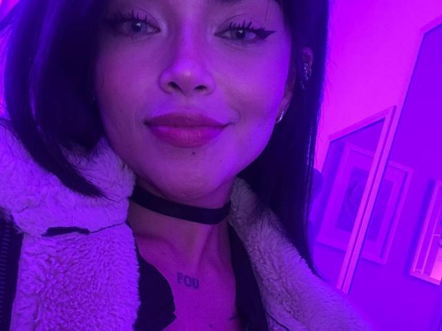 MajoJones with purple-lit face, black choker, white fluffy jacket, and red lipstick, showing small tits with tattoo "POU" above. She has long black hair and subtle smile.