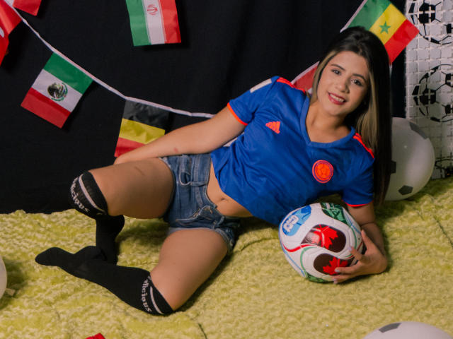 SheylaSpooner, with long brown hair, blue soccer shirt, red trim, denim shorts, black socks, holding soccer ball, lying on green carpet, smiling, with flags in background.