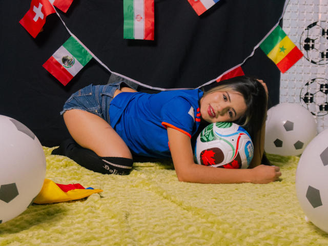 SheylaSpooner, with long black hair, red lipstick, and blue soccer jersey, lies on yellow carpet, butt up, knees to soccer ball, surrounded by soccer balls and flags.