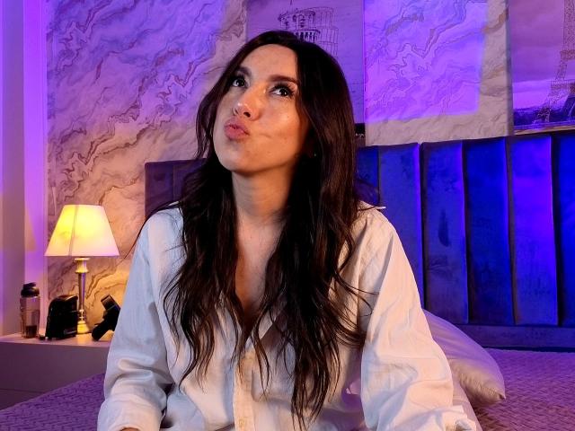 MiaReed has long brown hair, wearing a white shirt, pouting with her lips, sitting on a bed with blue and purple lighting. Bed has a blue headboard.