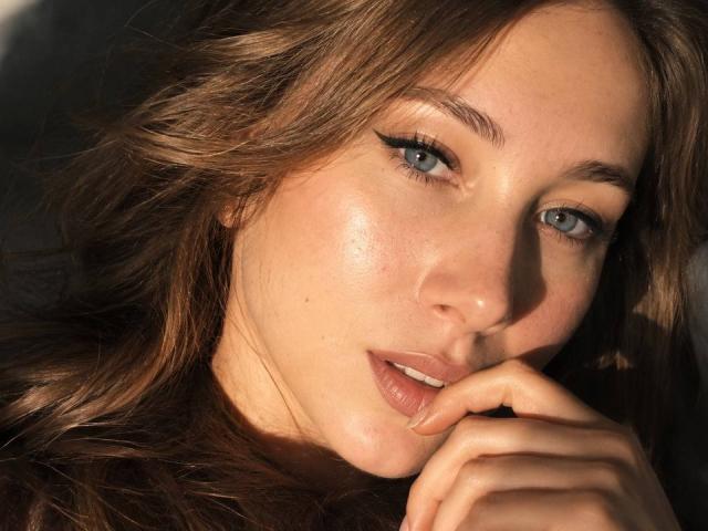 AmmaLoyXX has sexy blue eyes, full pink lips, and wavy brown hair, with a slightly shiny, freckled face, and her finger gently touching her lip.