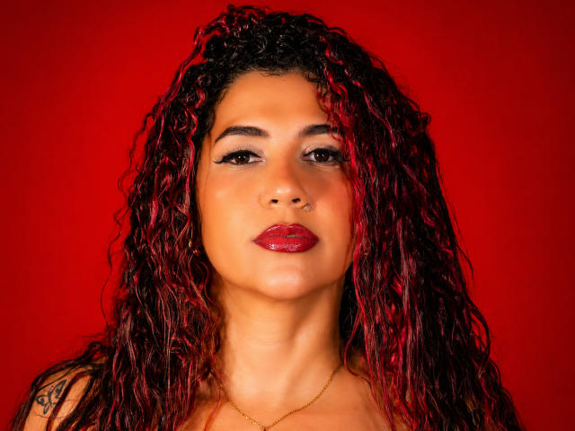 AntonelaRiveiro has red-pink curly hair, dark eyebrows, red lipstick, and a nose ring. She's wearing a gold necklace against a solid red background.