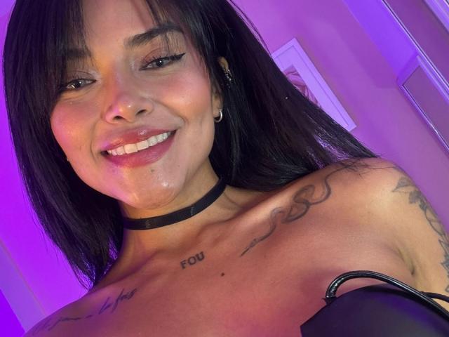 MajoJones with dark hair, smiling, black choker, tattoos, shiny wet skin, and visible chest tattoos, purple lighting, wearing black strap, looking at camera.