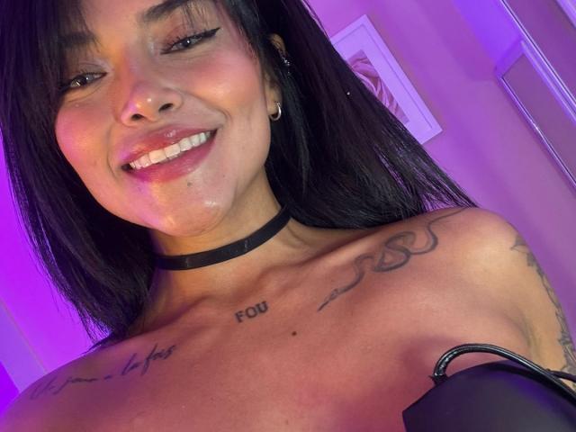 MajoJones with black hair, smiling, showing her tits, tattoos on chest, choker, nose piercing, and a small amount of cum on her face. Purple light in the background.