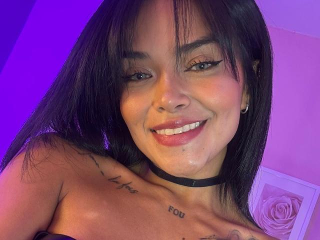 MajoJones with long black hair, smiling, wearing a black choker, showing tattoos on her shoulder and collarbone, purple and pink lighting, glossy skin, and cute nose.