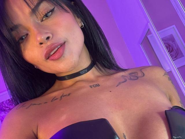 MajoJones with black hair, wearing a black choker, has tattoos on her chest and arms, and is topless with visible breasts in a purple-lit room.