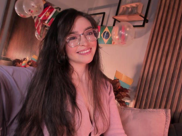 EvaMcAllister with long black hair, glasses, pink sweater, and a smile, showing her tits slightly, in a cozy, festive room with balloons and decorations.