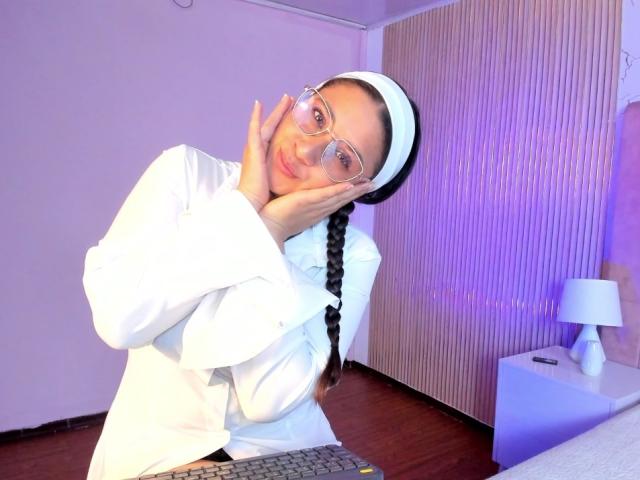 EmmyRossi with glasses, white headband, and braided hair, in a white shirt, resting chin on hand, smiling, in a room with purple lighting and a white lamp.