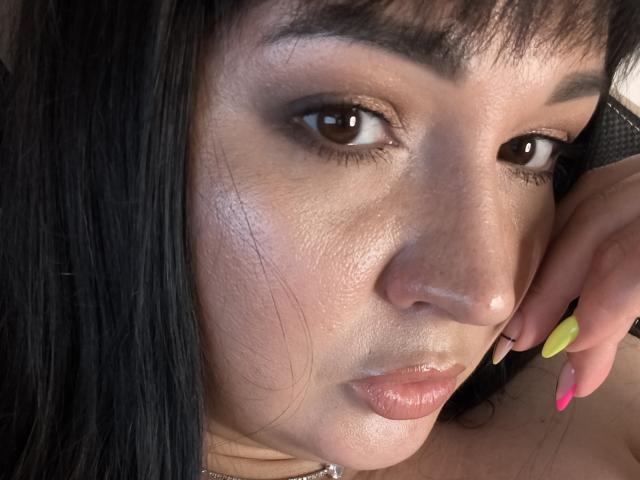 AlinaMilf's face, with shiny skin, brown eyes, and black hair, has a pouty expression and is resting her yellow and pink nail on her cheek.