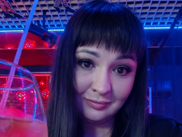 AlinaMilf with dark brown straight hair, bangs, and a slight smile, holding a drink with a straw in a blue and red neon-lit bar, showing her cute face.