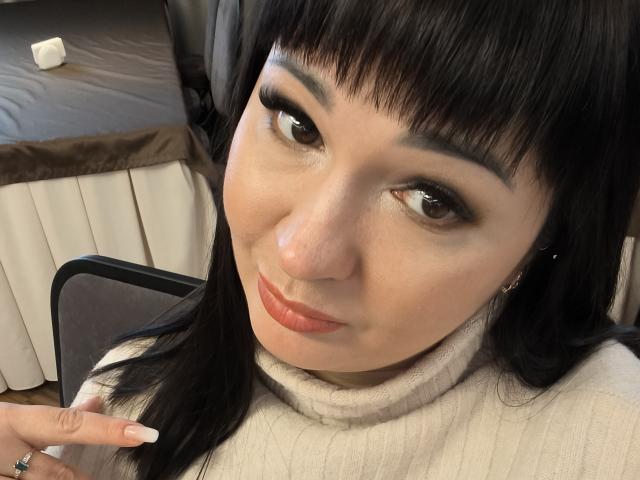 AlinaMilf has black hair, brown eyes, and is wearing a white turtleneck. She's touching her hair with manicured nails, smiling slightly with pink lipstick.