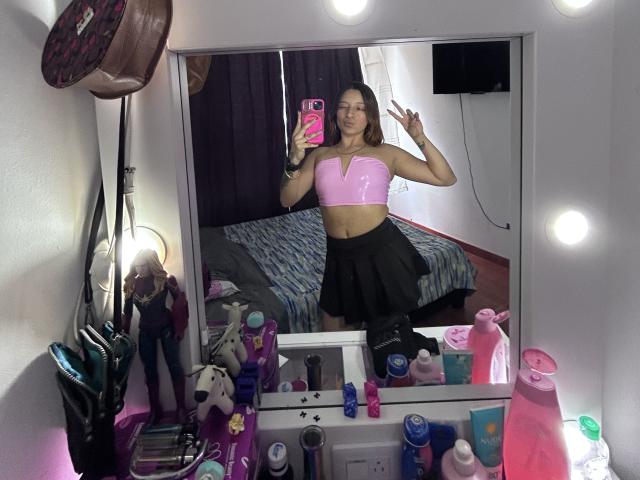 SaarahJones with mid-sized tits, wearing a pink shiny crop top and black skirt, making a peace sign, standing in front of a mirror with colorful makeup and toiletries.