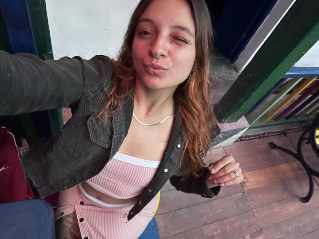 SaarahJones, pouty lips, winking, in a pink ribbed top, black jacket, blonde hair, wooden floor, green door, holding phone, ring on finger, casual, cute, small tits.