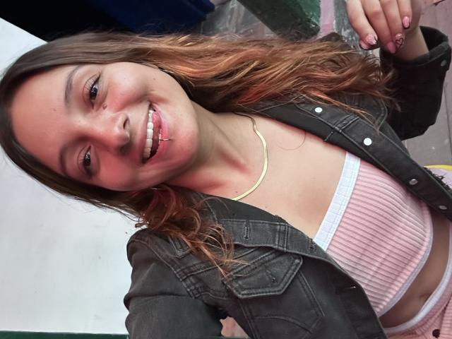 SaarahJones with wavy brown hair, smiling, wearing a black denim jacket, pink ribbed top, and white underwear, showing a small nose ring and a gold necklace.