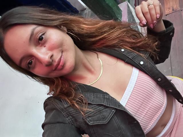 SaarahJones with wavy brown hair, wearing a pink ribbed crop top, white bra, black denim jacket, and pink high-waisted pants, smiling slightly, showing a nose ring and gold chain.