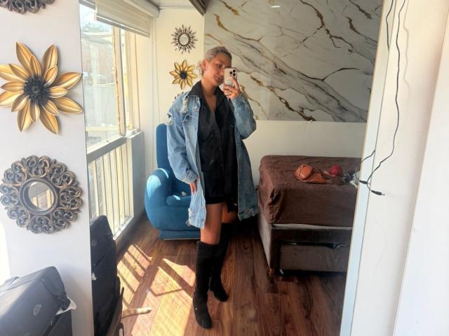 FloraSaenz in a blue denim jacket, black shorts, and thigh-high boots, holding a phone, standing in a room with marble walls, sunflower wall decor, and a blue chair.