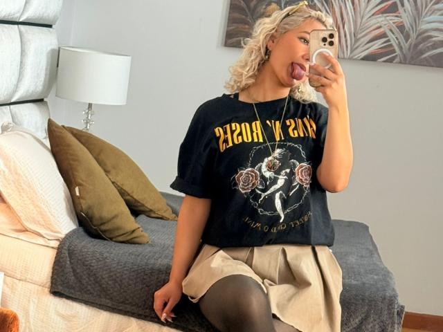 FloraSaenz with blonde curly hair, sticking out tongue, wearing black Nirvana shirt, beige skirt, black thigh-highs, sitting on bed, taking mirror selfie with iPhone.