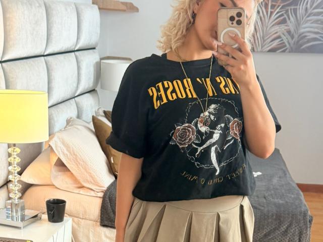 FloraSaenz with blonde curly hair, wearing a black "Iron Maiden" t-shirt and beige pleated skirt, taking a mirror selfie, showing her chest and holding a smartphone.
